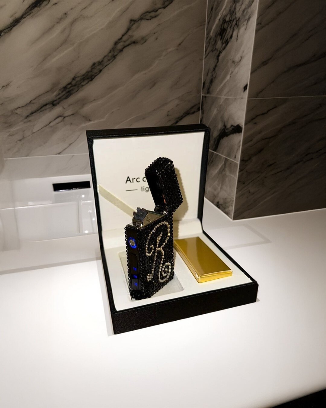 Crystal-covered arc lighter with a monogram, presented open in its black and white presentation box
